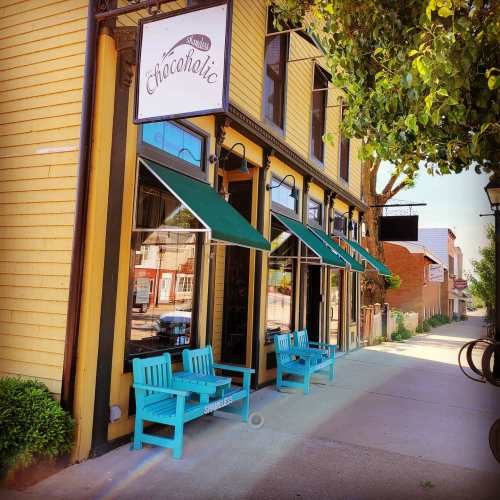 A charming storefront with teal chairs outside, featuring a sign that reads "Chocoholic" and green awnings.