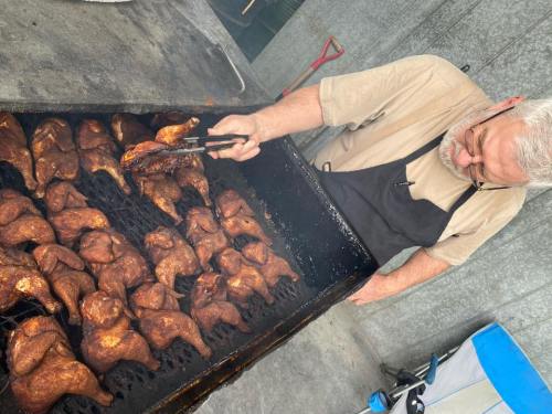 A man in an apron uses tongs to flip grilled chicken pieces on a large barbecue grill.