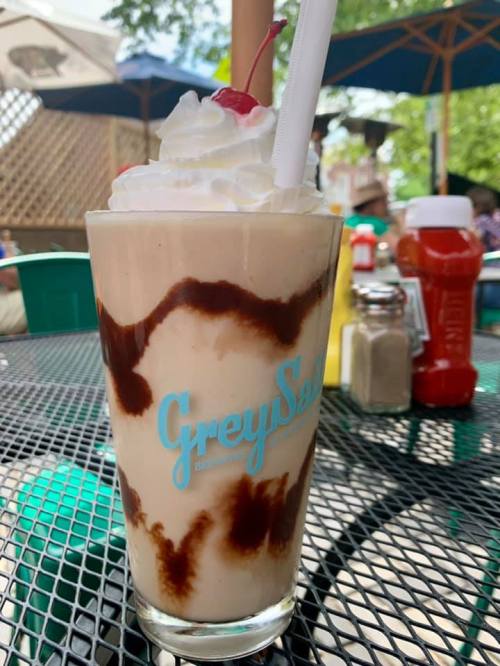 A tall glass of ice cream topped with whipped cream and a cherry, sitting on a table with condiments in the background.
