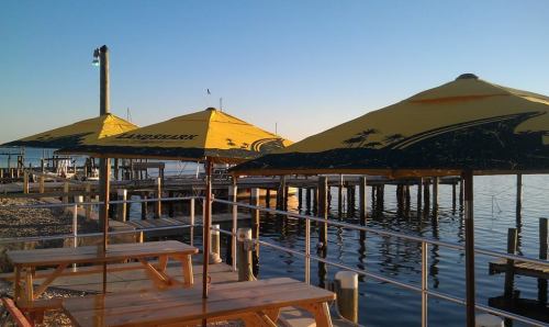 A waterfront scene with yellow umbrellas over wooden tables, reflecting calm water and a clear sky.