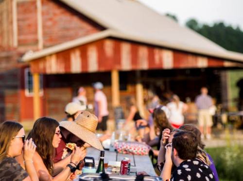 A group of people enjoying a meal at a long table outside a rustic barn during a sunny gathering.