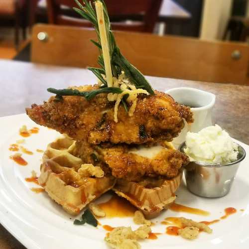 A plate featuring crispy fried chicken on waffles, drizzled with syrup, garnished with herbs, and served with whipped cream.