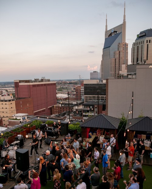 A lively rooftop event with a crowd enjoying live music against a city skyline at sunset.