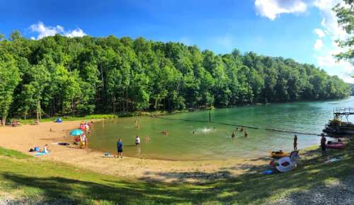 A scenic beach with people swimming and relaxing, surrounded by lush green trees and a calm lake under a blue sky.