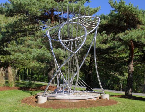 A modern metal sculpture with curved lines and vertical elements, surrounded by trees and greenery.
