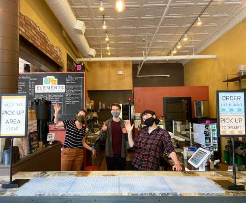 Three masked staff members wave from behind a counter in a café, with signs indicating order and pickup areas.