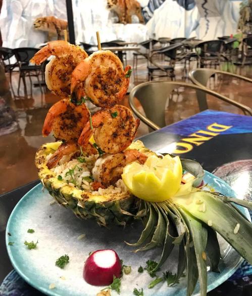 A vibrant dish featuring shrimp skewers atop rice served in a pineapple, garnished with lemon and herbs.