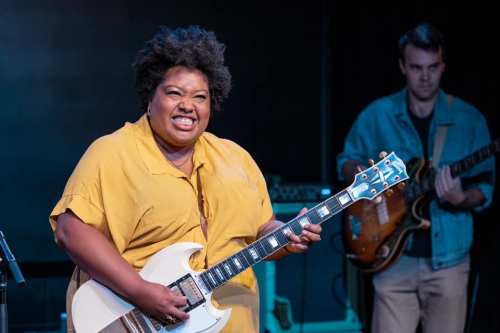 A smiling musician with an afro plays an electric guitar, while another musician plays bass in the background.