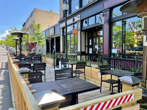 Outdoor dining area with wooden tables and chairs, surrounded by greenery, on a sunny street.