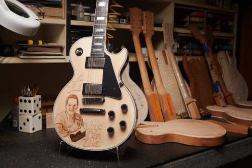 A custom guitar with an engraved portrait, surrounded by guitar bodies and tools in a workshop setting.