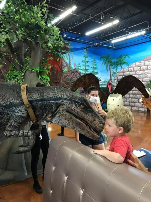 A child interacts with a lifelike dinosaur model in a colorful indoor setting, surrounded by other people and decorations.