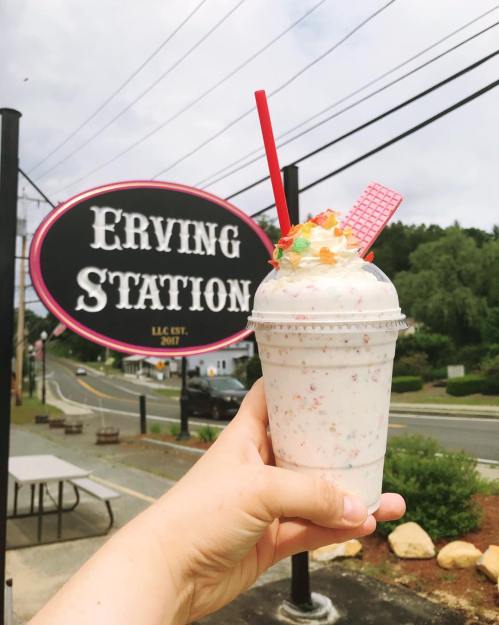A hand holds a colorful milkshake topped with candy, in front of a sign for Erving Station.