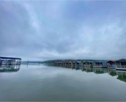 A calm lake reflects colorful boathouses under a cloudy sky, with distant hills in the background.