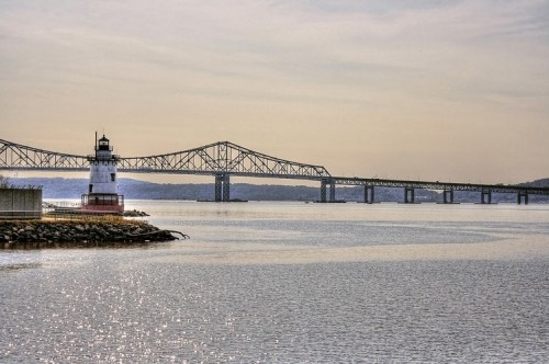 A lighthouse stands by the water, with a bridge spanning the river in the background under a clear sky.