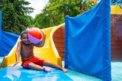 A smiling boy in red shorts holds a beach ball while sliding down a water slide in a colorful water park.