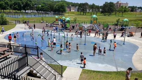 A busy splash pad with children playing in water, surrounded by playgrounds and green spaces on a sunny day.