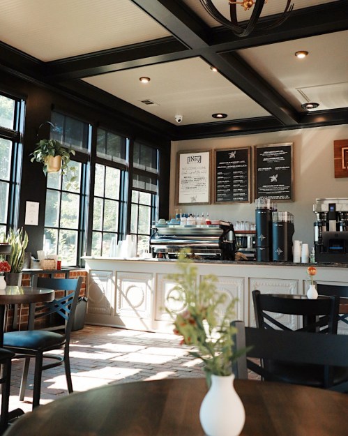 Cozy café interior with large windows, a coffee counter, and plants, featuring wooden tables and a warm atmosphere.