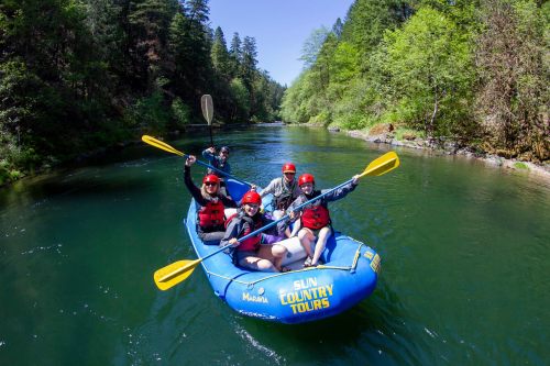 A group of five people in a blue raft, smiling and paddling on a serene river surrounded by lush greenery.