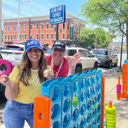 Two women wearing hats smile and pose in front of a colorful outdoor game near a tourist information sign.