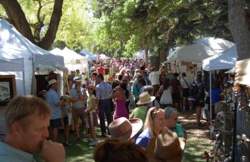 A bustling outdoor market with numerous tents, people browsing, and trees providing shade on a sunny day.