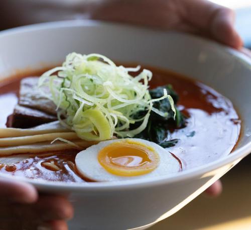 A bowl of ramen topped with sliced green onions, a soft-boiled egg, pork, and leafy greens, held by hands.