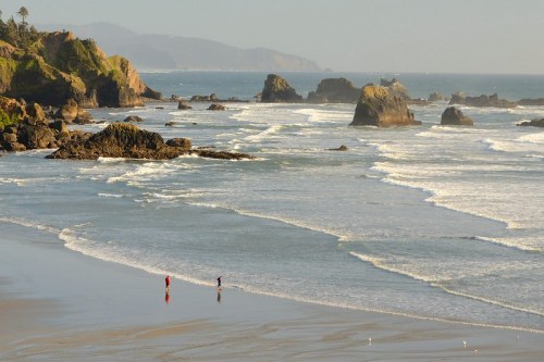 Two people walk along a sandy beach, with waves crashing and rocky formations in the background under a clear sky.