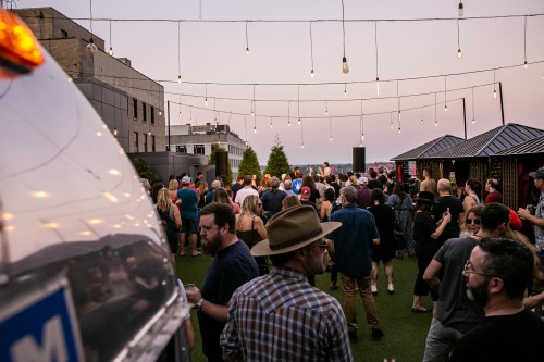 A lively rooftop event with people mingling under string lights, enjoying music and drinks as the sun sets.