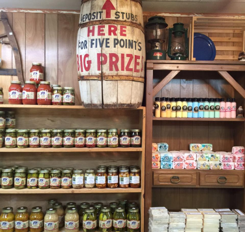 A rustic display of jars and soaps on wooden shelves, with a large barrel sign promoting a prize for deposit stubs.