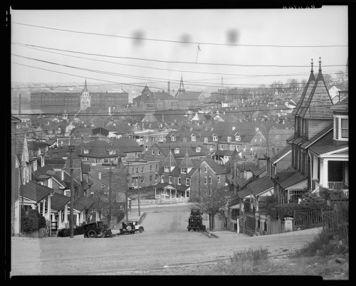 Black and white photo of a hilly neighborhood with houses, streets, and distant buildings under a cloudy sky.