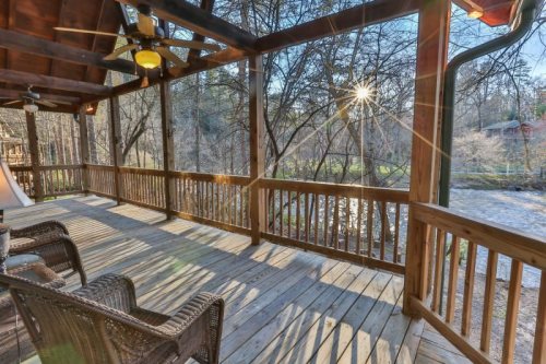 A wooden porch with two chairs overlooks a serene stream, surrounded by trees and sunlight filtering through.