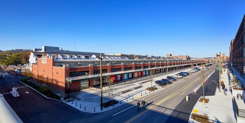 A sunny street view of a brick building with colorful signage, lined with parked cars and pedestrians.