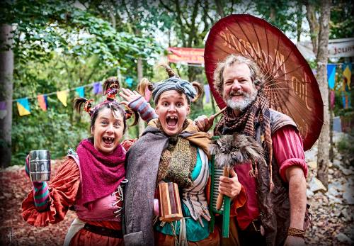 Three cheerful performers in colorful costumes pose together, smiling widely with props, surrounded by greenery and festive decorations.