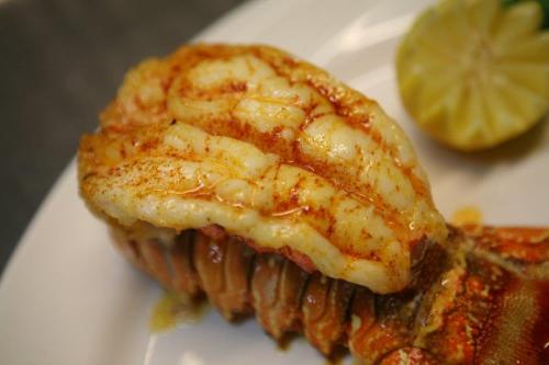 A close-up of a grilled lobster tail seasoned with spices, served with a wedge of lime on a white plate.