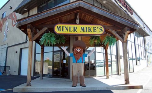 A large, friendly mascot with a beard and cowboy hat stands outside a building labeled "Miner Mike's."