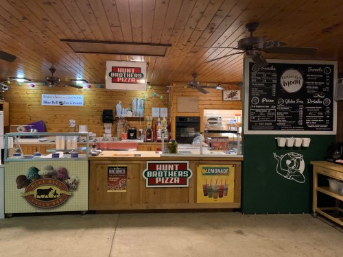 A rustic pizza counter with menu boards, ice cream display, and condiments in a cozy wooden setting.