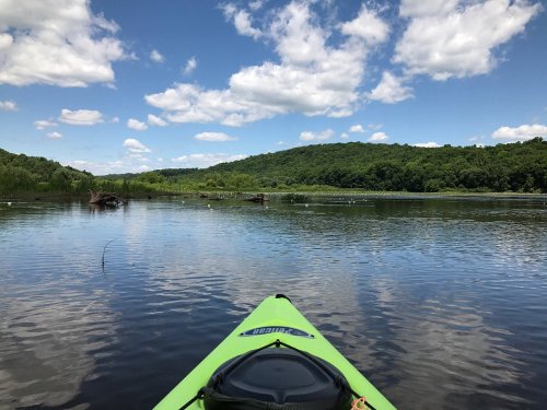 A kayak on calm water, surrounded by lush green hills and a blue sky with fluffy clouds.