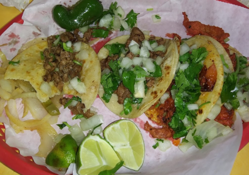 A colorful plate featuring three tacos topped with cilantro and onions, alongside lime wedges and jalapeños.