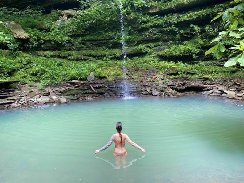 A person stands in a serene, turquoise pool with a waterfall and lush greenery surrounding them.