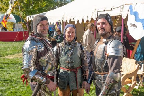 Three individuals in medieval armor pose together at a fair, with tents and people in the background.