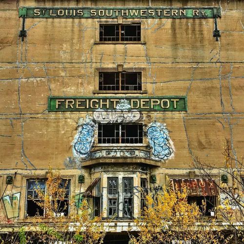 Abandoned freight depot building with faded signage and graffiti, surrounded by overgrown vegetation.