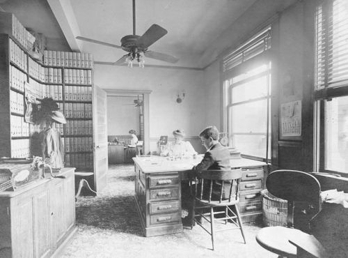 A vintage office scene with two people at a desk, shelves of files, and large windows letting in natural light.