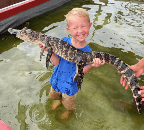 A smiling child stands in shallow water, holding a small crocodile with both hands.