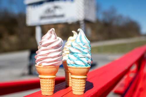 Three colorful soft serve ice cream cones in waffle cones, set against a bright outdoor background.