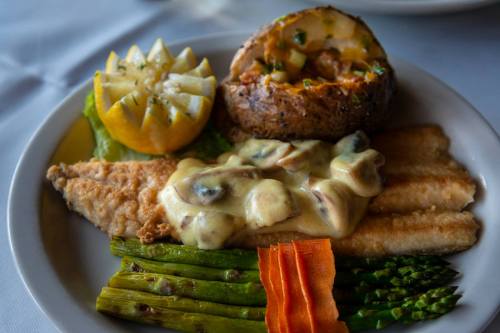 A plate featuring fried fish with creamy sauce, asparagus, carrots, a baked potato, and lemon wedges.