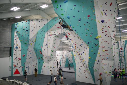 A spacious indoor climbing gym with colorful walls, climbers on various routes, and spectators below.