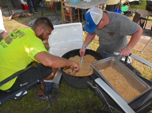 Two men cooking rice in a large pot outdoors, one stirring with a spoon while the other prepares a serving tray.
