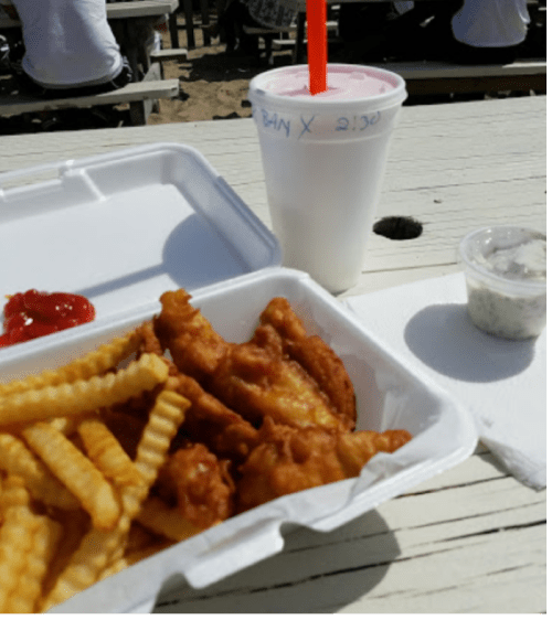 A takeout container with fries and fried fish, a cup of pink drink, and a small container of dipping sauce on a table.