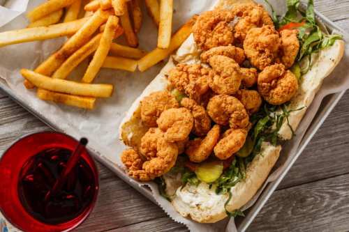 A sandwich filled with crispy fried shrimp, lettuce, and pickles, served with a side of golden French fries and a drink.