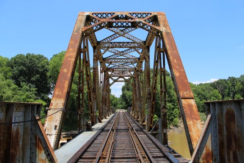 Rusty metal bridge with a wooden railway track, surrounded by green trees and a clear blue sky.