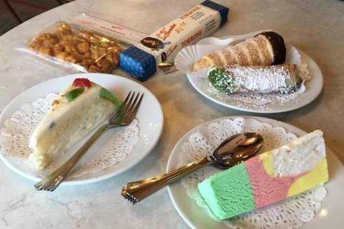 A variety of desserts on plates: cheesecake, cannoli, and colorful ice cream cake, with a box of torroncini in the background.